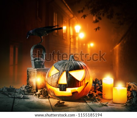 Scary horror background with halloween pumpkin jack o lantern, placed on wooden deck. Old town street on background with glowing lamps. Halloween spooky background.
