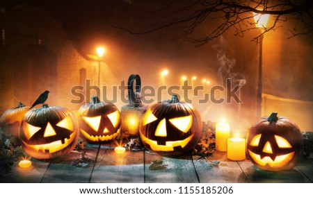 Scary horror background with halloween pumpkins jack o lantern, placed on wooden deck. Old town street on background with glowing lamps. Halloween spooky background.