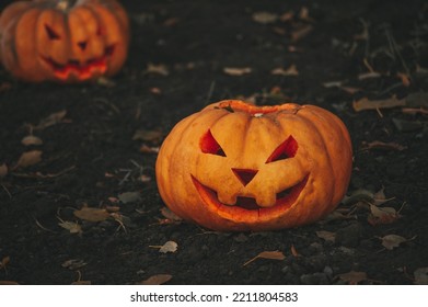 Scary Jack O Lantern Halloween Pumpkins In Sun Day On Ground Among Dry Leaves At Street. Hallows Eve Decoration Funny Glow Pumpkin With Candles On Candlestick At Fall Background In Open Air Near House