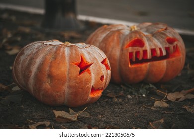 Scary Jack O Lantern Halloween Pumpkins In Sun Day On Ground Among Dry Leaves At Street. Hallows Eve Decoration Funny Glow Pumpkin With Candles On Candlestick At Fall Background In Open Air Near House