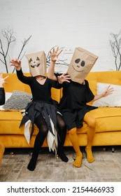 Scary Little Girls In Halloween Costumes Wearing Paper Bags With Scary Faces On Their Heads. Sitting On A Yellow Couch. Two Good Friends Having A Good Time.