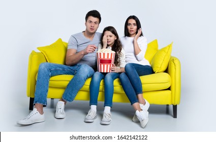 Scary movie! A small young family of three are sitting in the living room and watching horror films together with shocked facial expressions.