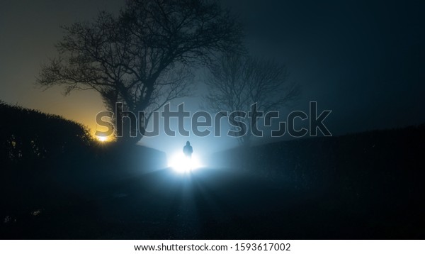 A scary, mysterious hooded figure, standing in\
front of a bright light on a country lane, on a spooky foggy\
winters night.