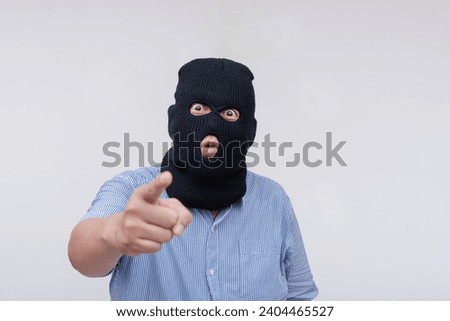 A scary and unhinged evil man wearing a black ski mask points at someone with his finger making threats. Isolated on a white backdrop.