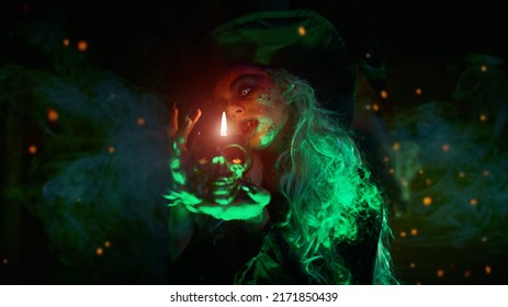 A Scary Wicked Witch Conjures In Her Lair And Holds A Skull Candle In Her Hands. Magical Green Light And Sparks Of Fire Around Her. Halloween. 