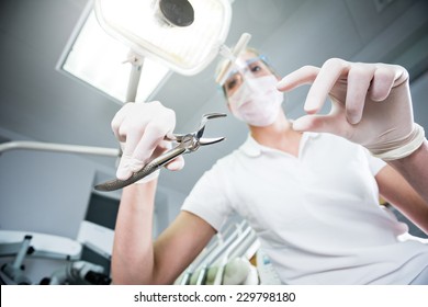 Scary Wide Shot Of Beautiful Doctor Inspecting Patient And Holding Pliers For Pulling Out The Tooth. Young Dentist In The Cabinet. View From Patient's Eye. Color Toned Image.