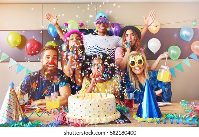 Scattered Confetti Falling Around Group At Party With Birthday Cake And Cone Hats On Table In Foreground