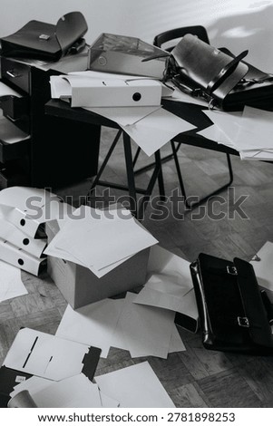 Scattered documents, folders, binders, and briefcases cover an office floor and desk, creating a cluttered workspace. Black and white tones, messy organization, paper chaos.