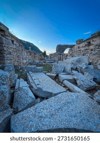 Scattered rectangular stone slabs and rubble dominate the foreground of Pergamon's Trajan Temple ruins, leading the eye through weathered walls to a prominent arched remnant. Distant mountains pierce a vivid blue sky under soft twilight illumination, highlighting textured limestone against sparse greenery.