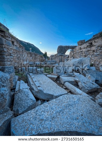 Scattered rectangular stone slabs and rubble dominate the foreground of Pergamon's Trajan Temple ruins, leading the eye through weathered walls to a prominent arched remnant. Distant mountains pierce a vivid blue sky under soft twilight illumination, highlighting textured limestone against sparse greenery.