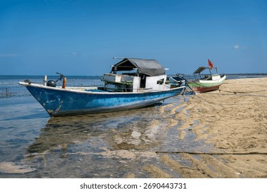 A scene featuring two rustic, traditional Indonesian fishing boats (jukung or similar) pulled up onto a sandy beach at low tide. The main boat is painted blue and white and shows signs of wear