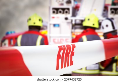 Scene of incident is cordoned off with red and white tape, reading ‘fire’. Out of focus in the background are three firefighters, two fire engine appliances and smoke billowing through the air.