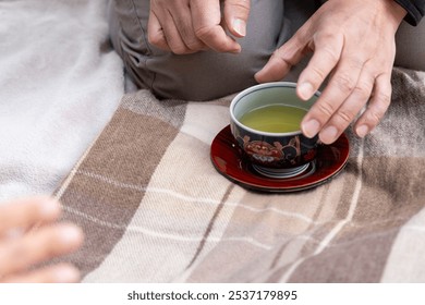 A scene from a Japanese autumn picnic in a park: a man's hand reaching forward to grab a cup of hot green tea, set on top of a brown and beige plaid scarf, and light pink picnic blanket. - Powered by Shutterstock