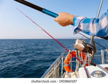 Scene on a sailboat in the Mediterranean. Several fishing rods are mounted. In the foreground a man in a shirt is holding a fishing rod. Close up of the rod and reel. Sunshine and blue water.