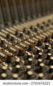 The scene shows a detailed view of knobs and sliders on an audio mixer in a recording studio. The lighting is dim, typical of a music production setting.