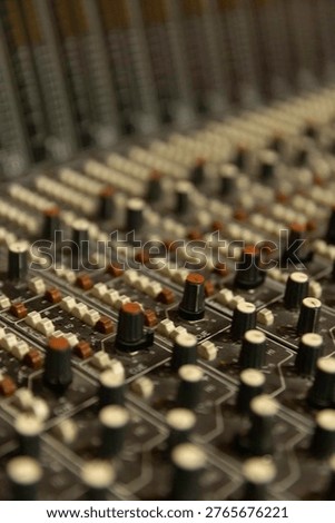 The scene shows a detailed view of knobs and sliders on an audio mixer in a recording studio. The lighting is dim, typical of a music production setting.