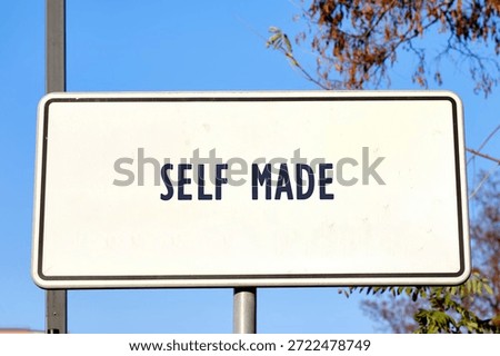 The scene shows a rectangular white sign with dark lettering that proclaims SELF MADE. It is attached to a metal post with bare tree branches and a vast blue sky in the background.