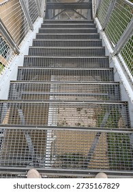 The scene shows a view down a long mesh staircase from a walkway that crosses over a busy highway. The metal stairs are clearly visible with a view of the ground below.