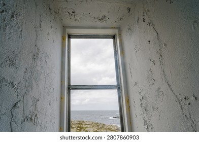 The scene shows a window frame inside Vinga Lighthouse. Outside, the sea meets rocky land under a cloudy sky. The waves gently break on the shore.