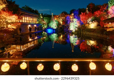 Scenery of beautiful autumn foliage illuminated at blue dusk and reflected in the water of a pond with small lanterns tied under bamboo railing in Mejiro Garden (目白庭園) in Toshima Ku, Tokyo City, Japan