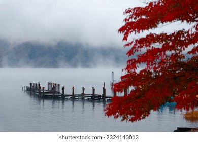 Scenery of beautiful Lake Chuzenji 中禅寺湖 on a foggy autumn day, with wooden piers extending onto the misty water and fiery maple foliage on the lakeside, in Nikko National Park, Tochigi, Japan