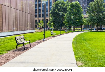 Scenery Of Bench Placed Along Walkway In Green Public Park, Lush Grass Around Outdoor Garden For Recreation And Rest In Sunny Day. Spring And Summer Lifestyle With Nature Environment. Empty Landscape.