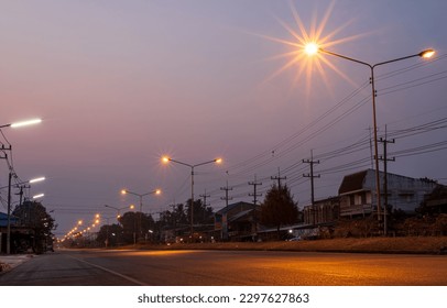 Scenery of orange starbursts from street lamp posts illuminating the road near residential houses and electric poles in rural Thailand with early morning sky in the background.