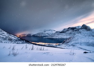 Scenery of snow covered mountain range with illuminated town on coastline in blizzard at mount Segla, Senja island, Norway