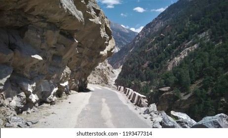 Scenery of the state highway at Spiti Valley, India. A barren rocky mountain forms a semi-arch over the road, while trees cover the opposite mountain. Boulders and concrete kerbs form the side barrier