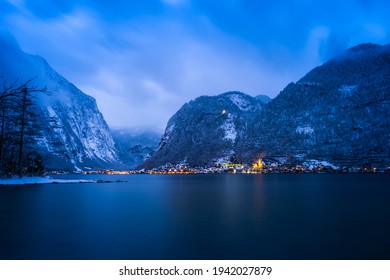 Scenery View Of Austrian Snowy Village Hallstatt By Lake Surrounded By Snow Mountains With Lights On After Sunset At Night Time, Austria. Image Contains Of Noise And Dust From Snow Falling Down.