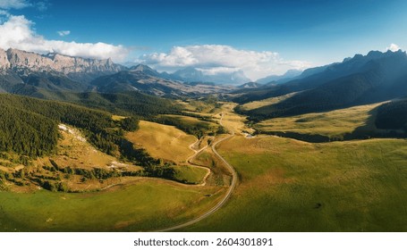 A scenic aerial shot captures a winding road traversing a lush green valley nestled between majestic mountain ranges under a vibrant blue sky. Clouds softly caress the peaks.