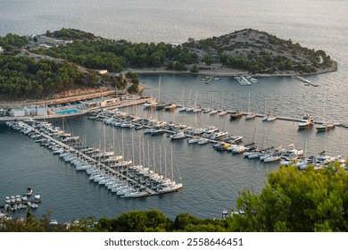 Scenic aerial view of marina with numerous sailboats and yachts docked in surrounded by lush green hills and rocky coastline at sunset. Maritime tourism, sailing adventures, and coastal relaxation. - Powered by Shutterstock