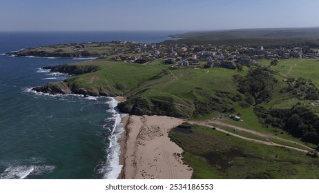 A scenic aerial view of a rugged coastline featuring a solitary house on a cliff, surrounded by crashing waves. The coastal town in the distance adds charm to the natural beauty of the rocky shore and - Powered by Shutterstock