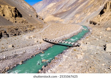 A scenic aerial view of a small stone bridge crossing a turquoise river, set against a backdrop of towering, barren mountains. The rugged landscape and pristine water create a dramatic - Powered by Shutterstock