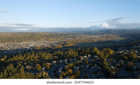  A scenic aerial view of a suburban neighborhood surrounded by autumn-colored trees and open countryside, leading to the iconic snow-capped Mount Rainier in the distance. - Powered by Shutterstock