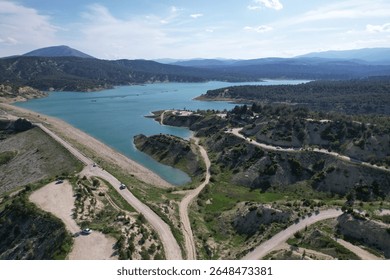 Scenic aerial view of a winding road along a tranquil turquoise lake surrounded by green hills and pine forests. The distant mountain range under a blue sky adds depth and serenity to the landscape. P