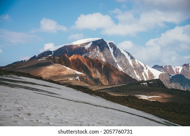 Scenic alpine landscape with snow-capped mountain peak and vivid red sharp rocks under blue sky with clouds. Colorful sunny mountain scenery with snow mountain top and pointy motley orange rocks.