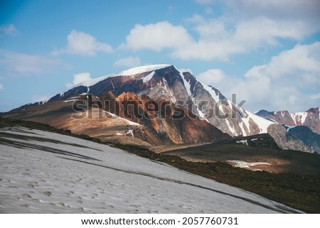 Scenic alpine landscape with snow-capped mountain peak and vivid red sharp rocks under blue sky with clouds. Colorful sunny mountain scenery with snow mountain top and pointy motley orange rocks.