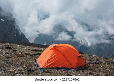 Scenic alpine landscape with vivid orange tent at very high altitude with view to high snowy mountain and large glacier in dense low clouds. Awesome mountain scenery with tent in thick low clouds.