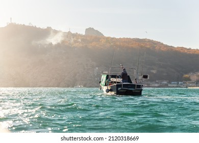 Scenic Back View Of Fisherman Angler Enjoy Fishing With Rods On Motor Boat Ship At Clean Green Blue Azure Water Harbor On Early Misty Morning Sunrise. Fishing Speedboat With Angling Man At Foggy Bay