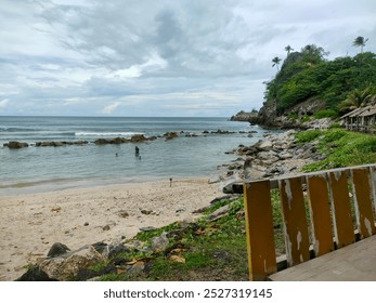 Scenic beach with rocky shoreline, calm ocean waves, and lush green hills under a cloudy sky, creating peaceful vibes.

 - Powered by Shutterstock