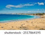 A scenic beach view with golden sand and clear turquoise waters. People are enjoying various activities on the beach, including swimming and sunbathing. At Towan Beach, Newquay, Cornwall, England.