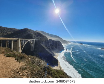 The scenic Bixby Creek Bridge on California's Highway 1, with dramatic coastal cliffs, a sunlit sky, and the Pacific Ocean's waves crashing below. The sun creates a striking lens flare. - Powered by Shutterstock