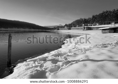 Scenic black and white image of lake / river snow covered bank, with old, weathered water level measuring pole, reflective water surface, metal barrels supported surface and hills in the background 