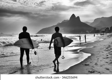 Scenic Black And White View Of Rio De Janeiro, Brazil With Brazilian Surfers Walking Along The Shore Of Ipanema Beach