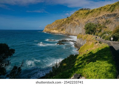 Scenic coastal landscape with dramatic cliffs, a winding coastal road, and waves crashing against the rocky shore under a clear blue sky. The vibrant greenery in the foreground contrasts - Powered by Shutterstock