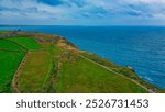 A scenic coastal landscape featuring lush green fields, a winding path, and a dramatic view of the ocean under a cloudy sky. In Tintagel, Cornwall, UK.