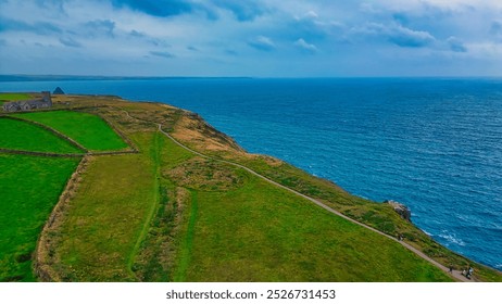 A scenic coastal landscape featuring lush green fields, a winding path, and a dramatic view of the ocean under a cloudy sky. In Tintagel, Cornwall, UK. - Powered by Shutterstock