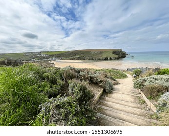 A scenic coastal path with wooden steps leading to a sandy beach, surrounded by lush greenery, cliffs, and calm blue waters under a partly cloudy sky. Houses dot the distant hillside. - Powered by Shutterstock