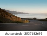 Scenic coastal road with a sharp curve, overlooking a serene sea and mountains in the distance during sunset, creating a calm and peaceful ambiance.